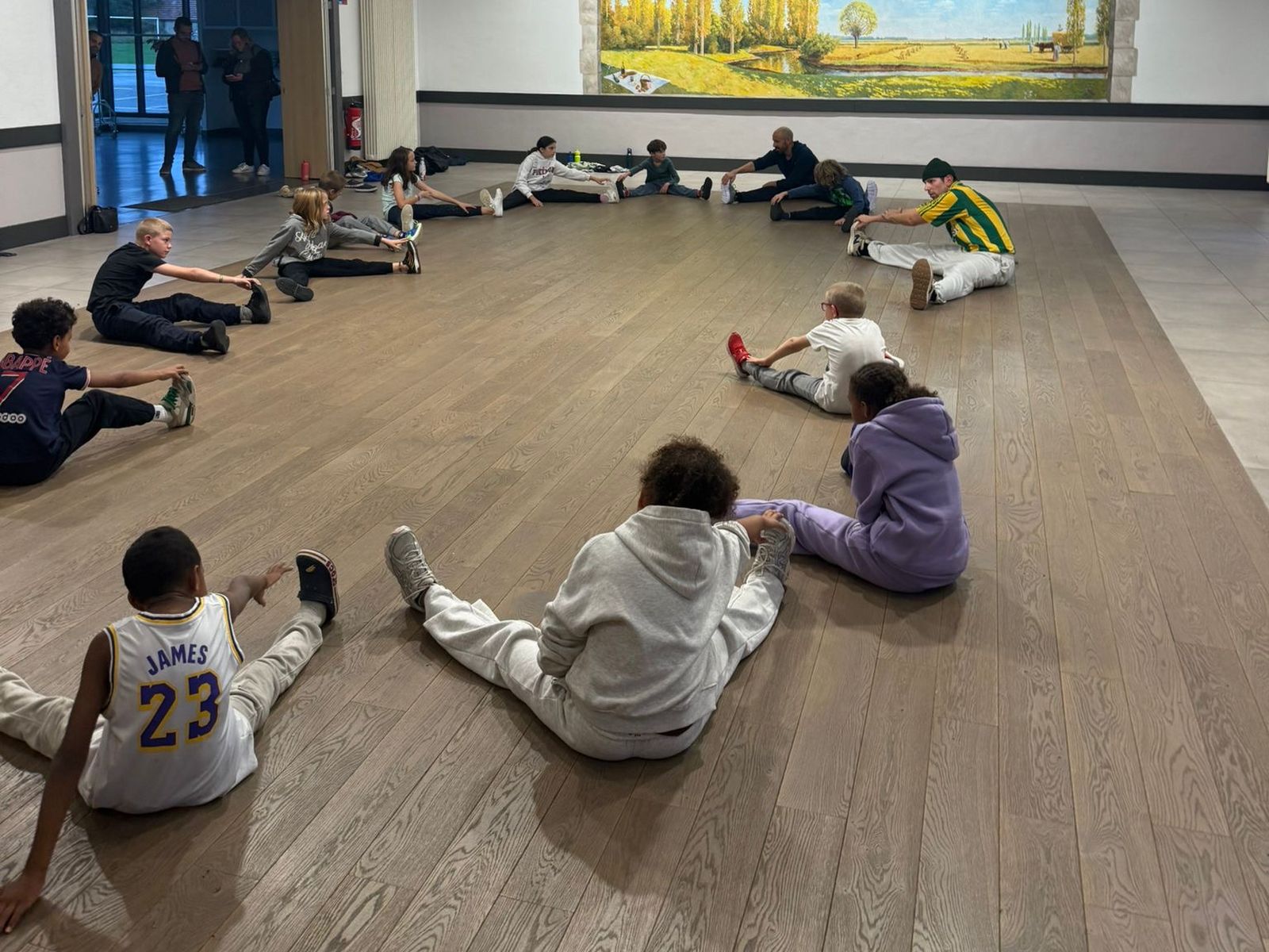 Les élèves assis en cercle sur le parquet font des étirements jambes écartées